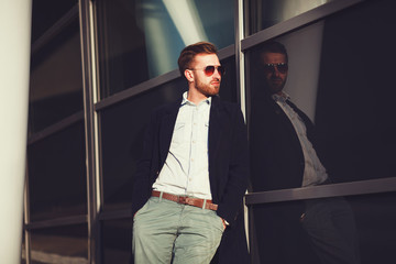 Young handsome businessman posing leaning against a glass, wearing a coat - business concept