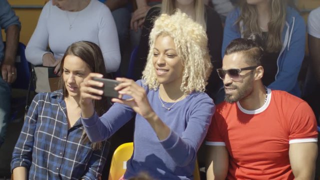  Friends sitting in the crowd at sports event taking photos with smartphone
