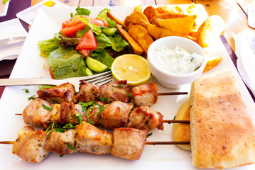 Traditional Cyprus souvlaki on white plate with salad, potatoes, bread and tzatziki sauce.