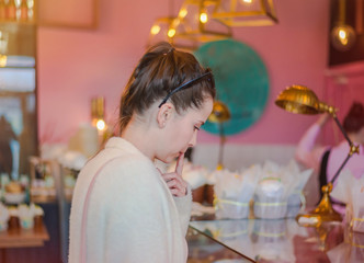 Beautiful brunette girl in elegant interior cafe, choosing some sweets to buy. Pretty young woman in cozy comfortable cafe going to make an order. Selective focus.