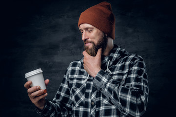 A brutal bearded male dressed in a hat and fleece shirt, drinks coffee from a paper glass.