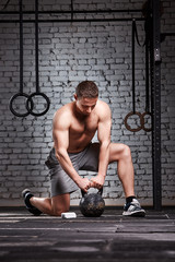 Young sportsman holding kettlebell with his arms on the cross fit gym floor against brick wall.