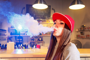 Young pretty woman in red cap smoke an electronic cigarette at the vape shop