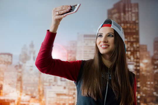A young and positive girl is photographed on her smartphone in her room on the background of the city depicted on the wallpaper.