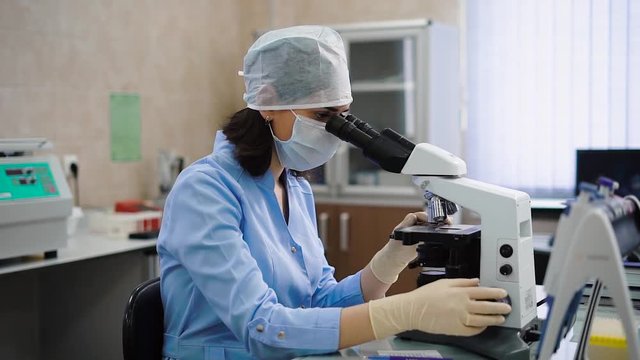 Female laboratory worker using microscope. Young female specialist in uniform sitting at table and using microscope.