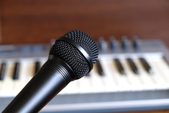 Black vocal microphone close up against defocus electronic synthesizer keyboard with many control knobs in silver plastic body as background
