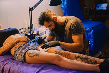 Fototapeta na wymiar The bearded tattoo artist makes a tattoo on a woman's leg in an ink saloon.