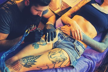 The bearded tattoo artist makes a tattoo on a woman's leg in an ink saloon.