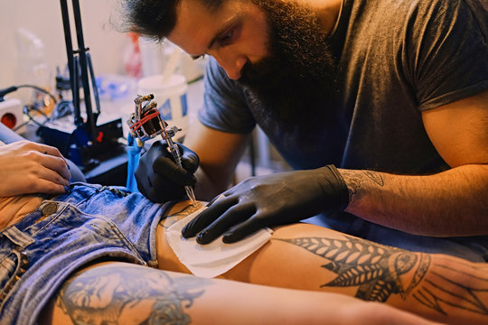 Bearded tattoo male artist makes a tattoo on a female leg.