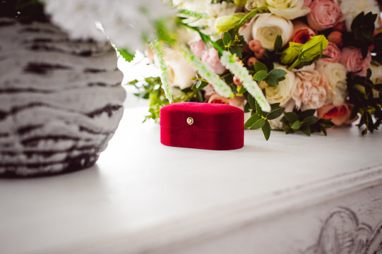 Photo image of a red velvet box with wedding rings of the bride and groom, on a white table, with a bridal bouquet of bridal flowers