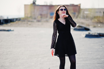 Portrait brunette girl with red lips and orange mobile phone at hands, wearing a black dress, sunglasses posed on the roof. Street fashion model.