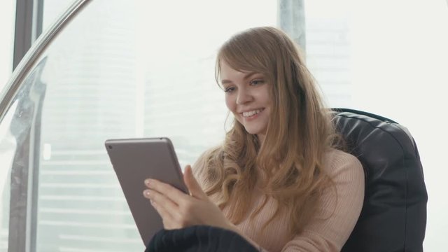 Young woman at home sitting on modern chair in front of window and working with tablet computer