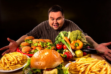 Diet fat man who makes choice between healthy and unhealthy food . Overweight male with hamburgers, french fries and vegetables trays trying to lose weight first time .