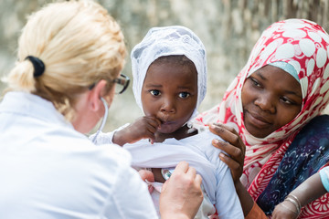 Female Caucasian doctor listening heart beat and breathing of little African girl with...