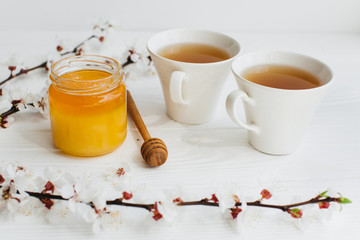 Bank of honey, spoon, blossom branches and two cups of hot tea on a white wooden background.