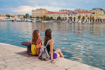 Two young girls with backpacks sitting on the stone pavement near the sea shore looking at the marine and pretty little colorful houses. Town of Zadar, Croatia - obrazy, fototapety, plakaty