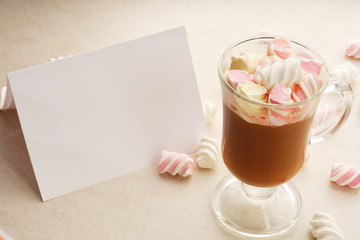 A blank sheet of paper, a glass of hot chocolate with marshmallows on a light background.