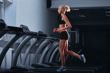 Horizontal full length shot of an attractive fit young woman with perfect strong muscular body wearing sports top and shorts jogging on a treadmill at the health club diet slimming toning motivation . © serhiibobyk
