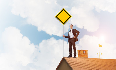 Young businessman on house brick roof holding yellow signboard. Mixed media