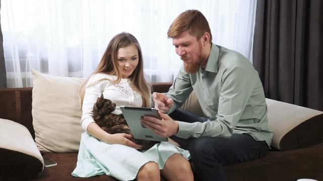 Couple is sitting on the couch sofa at home Look into the tablet and smile, Drink tea from a white tea set and watching tv.