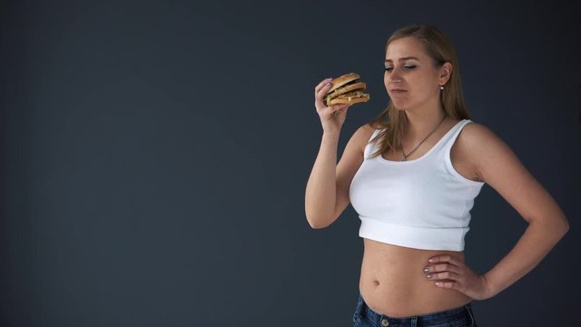 Overweight woman with a hamburger on a gray background. She thinks or eat fast food