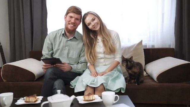 Couple A young man and a woman, and their cat is sitting on the couch sofa at home Look into the tablet and smile