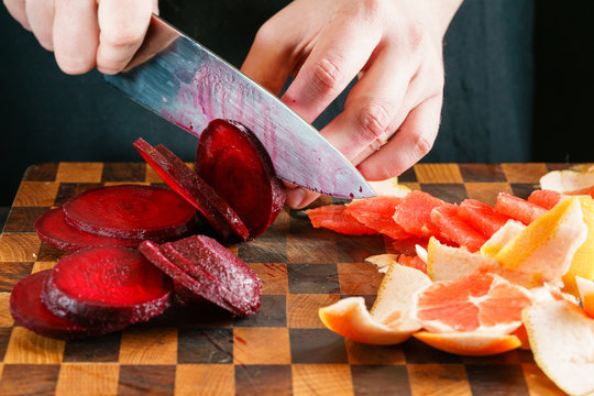 Chef's Hands Slicing Red Beet With A Large Kitchen Knife On A Wooden Cutting Board. Nearby Is Already Peeled And Sliced Grapefruit Or Orange.