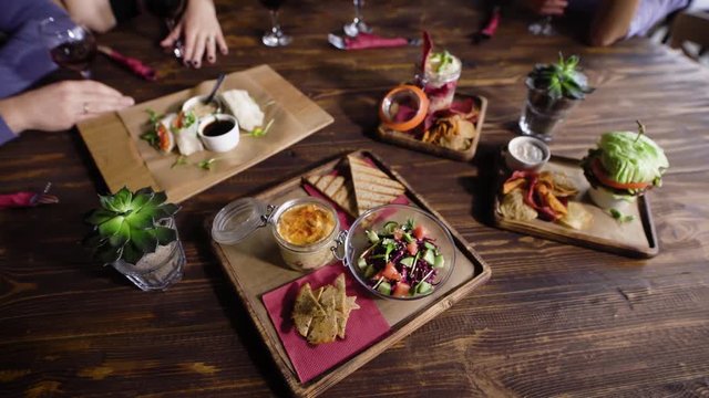 Close up of table served with assorted food setting on wooden platters. Image of different dishes and snacks on the brown surface with people and glasses of wine in the background.