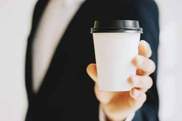 Closeup view of Businessman holding white paper coffee cup to take away.Mock up of carton coffee, cup for go outside.Horizontal mockup, blurred background.