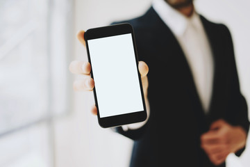 Businessman in a black suit showing smartphone with vertical blank white display.Horizontal mockup, blurred background.