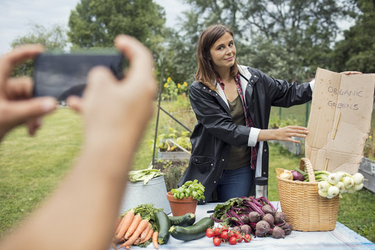 Cropped image of man photographing woman posing at table full of garden vegetables