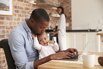 Father and baby sitting at laptop while mother prepares meal