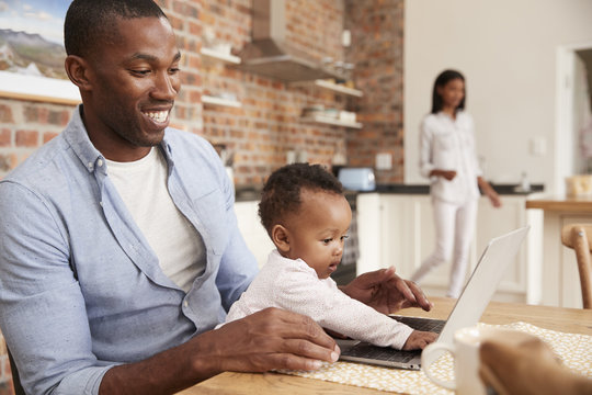 Father And Baby Daughter Use Laptop As Mother Prepares Meal