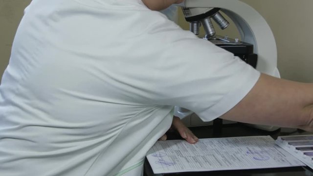 Middle aged caucasian female lab technician in white uniform working with microscope at laboratory, tilt up from hands to portrait of woman, concept medical analysis and diagnosis