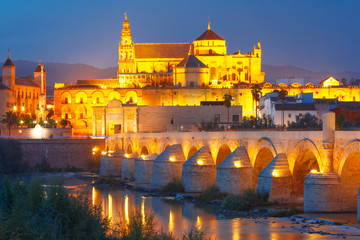Illuminated Great Mosque Mezquita - Catedral de Cordoba with mirror reflection and Roman bridge across Guadalquivir river during evening blue hour, Cordoba, Andalusia, Spain