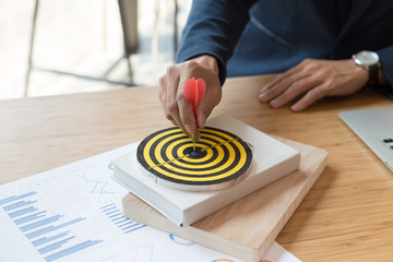 Businessman 's hand holding dart hitting at the business target on dartboard on desk in office. ...
