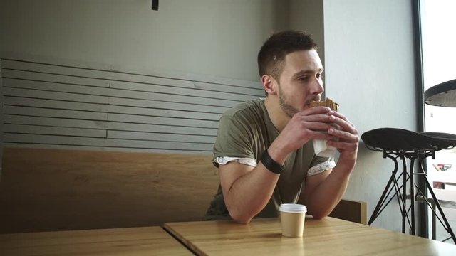 Young handsome Caucasian male in olive t-shirt sitting at cafe eating sandwich in slowmotion