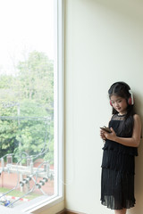 Pretty asian girl holding smart phone and listening to music with her headphones near window