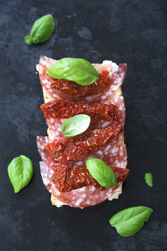 Simple sandwich with salami, basil and dried tomatoes on a dark textural background