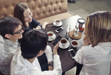 Group of young people from two couples of guys and girls have a tea time in the cafe and looking on the smartphone absorbedly