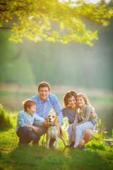 A large family is walking his beagle in a park by the lake. Mom, Dad, son and daughter are sitting on the grass under a tree on a warm summer evening