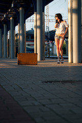 Lonely young brunette woman, in sexy shorts with old suitcase on station platform with sunlight