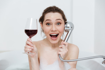 Cheerful woman drinking wine and joking using shower in bathtub