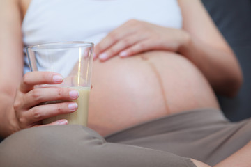 Beautiful pregnant woman holding a glass of milk and keeping a hand on her bare tummy while sitting in the living room