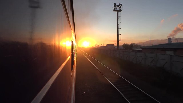 Passenger train goes by railway to the orange sun. Sunrize sunset morning. Locomotive From the window FOV, reflection. Populated aerial city at horizon. Dust industry smoke