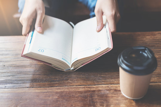 Focus on book. Woman holding notebook with a cup of coffee on wooden table.