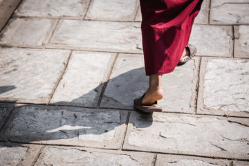 Feet ot a buddhist monk wearing sandals and traditional red robe walking in the midday sun with a...