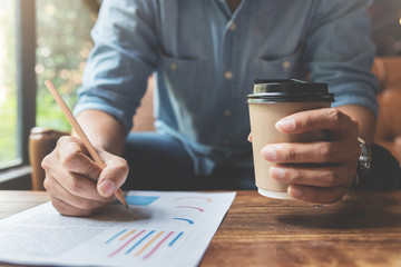 Close up Freelance or business man hands holding cup of coffee and writing on document with pencil. Graph financial diagram documents on wooden table.