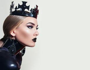 Black queen. A stern look. Black crown. A princess. Maleficent. Girl with a black crown decorated with precious stones sitting in studio on light background. Dark lipstick.