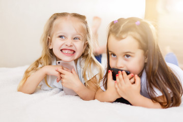 people, children, technology, friends and friendship concept - happy little girls with smartphones lying on floor at home education, school are using a smartphone and smiling while sitting on sofa